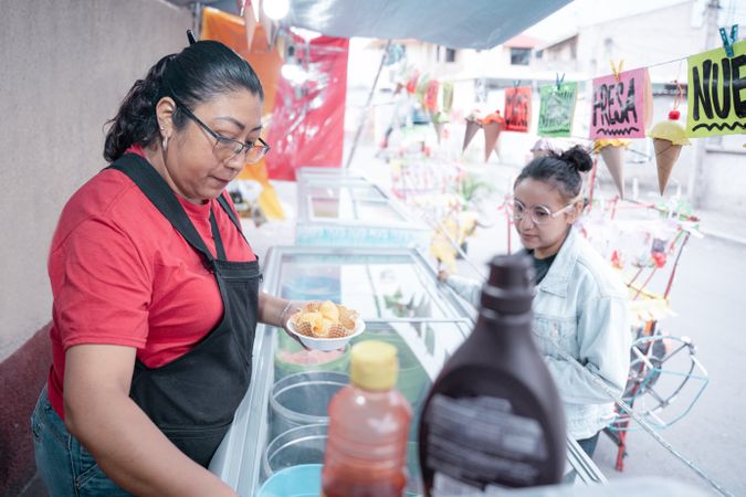 An adult Hispanic woman is preparing a vanilla ice cream for a young woman client in a street market stall