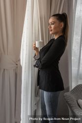 Side view of woman in dark blazer drinking coffee standing by the window 4jM6X5