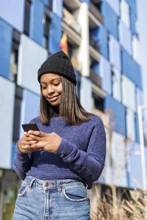 Female in hat and blue sweater checking cell phone outside blue building, vertical