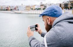Guy on vacation in casual clothing making photo of bay on smartphone at quay