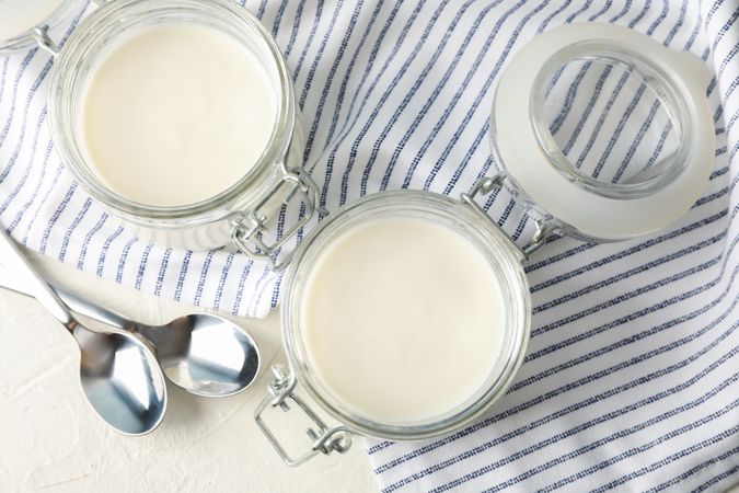 Glass jars with sour cream yogurt, spoons and towel on cement background
