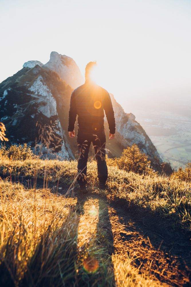 Back view silhouette of a man standing on brown grass facing the sun and mountains