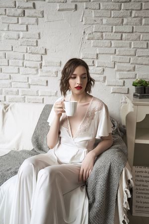 Pensive adult woman looking away and dreaming while sitting on sofa and having hot drink in light modern living room