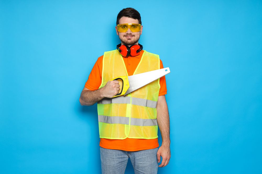 Worker with saw in goggles and protective ear muffs on blue background
