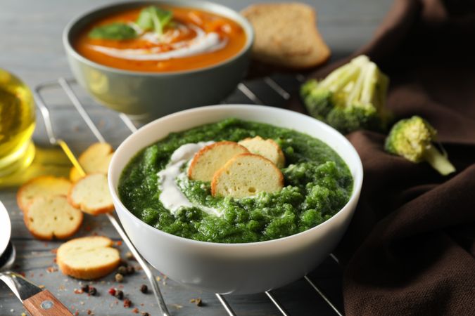 Two bowls of orange and green soup on wooden table with crackers and vegetables