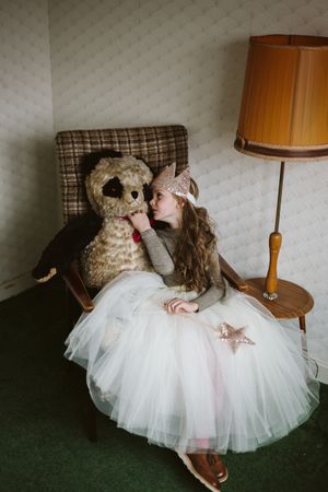 Young girl in light dress sitting on armchair holding bear plush toy