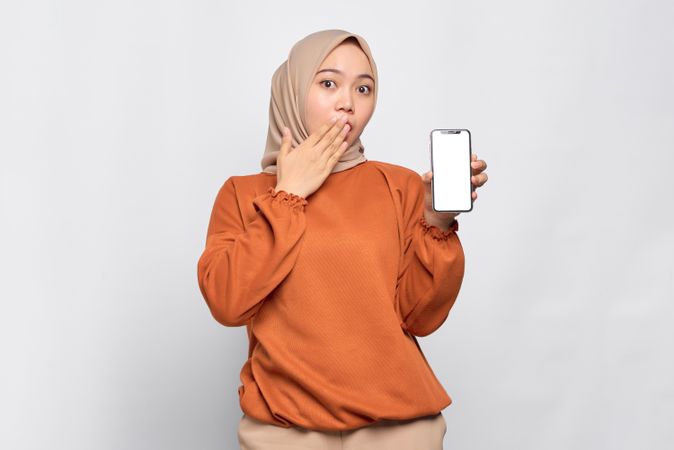 Amazed Muslim woman smiling with hand over mouth presenting smart phone with mock up screen