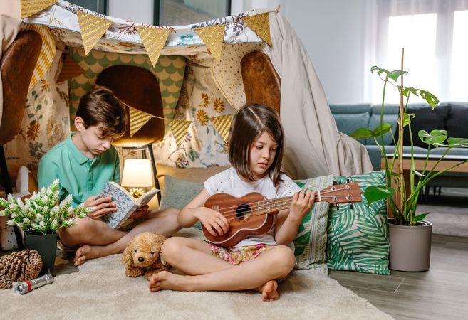 Little girl playing ukulele while boy reading book on handmade teepee in living room at home