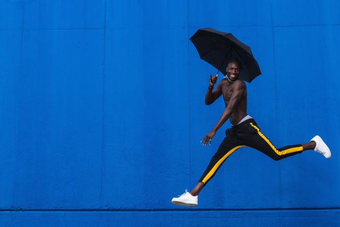 Cheerful Black man with umbrella jumping on street