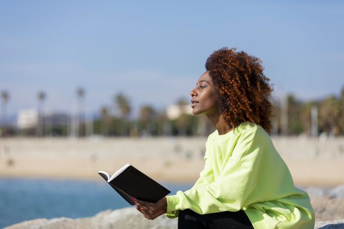 Content female in bright green shirt relaxing on the seaside on sunny day with a book