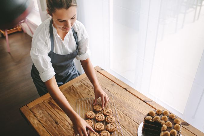 Female baker arranging cookies on rack with cake on table