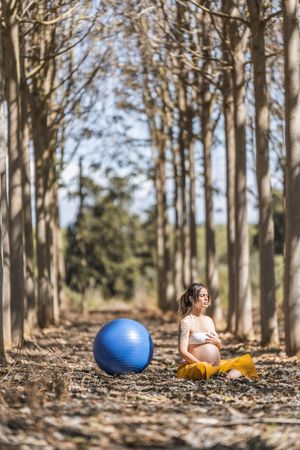 Serene adult pregnant woman meditating while sitting on ground among trees in park during sunny day