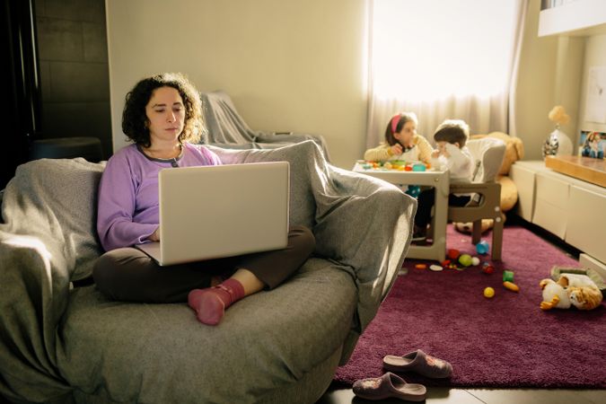 Mother working on her laptop sitting in living room while a girl and boy playing