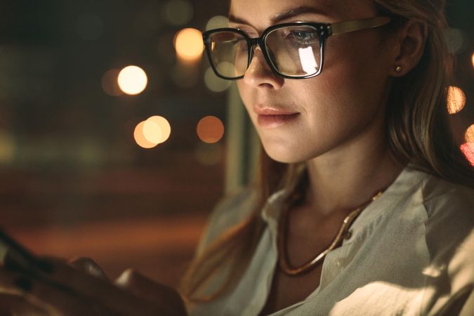 Close up of young woman wearing glasses using cellphone in office at night