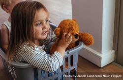 Beautiful little girl sitting in a washing basket with her teddy bear and brother at the back 5rOKM4