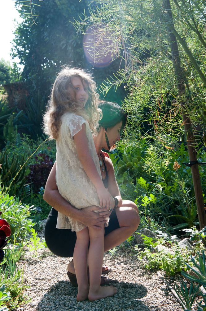 Mother puts arm around  daughter on rock path looking at plants with child looking at camera