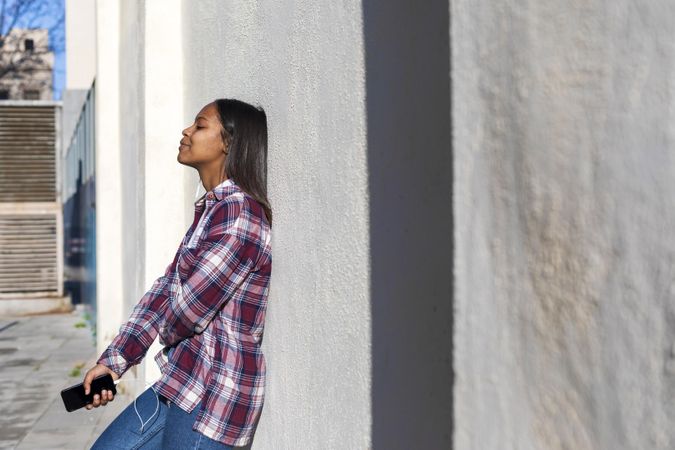 Female standing in the sun while leaning on wall listening to music on smartphone