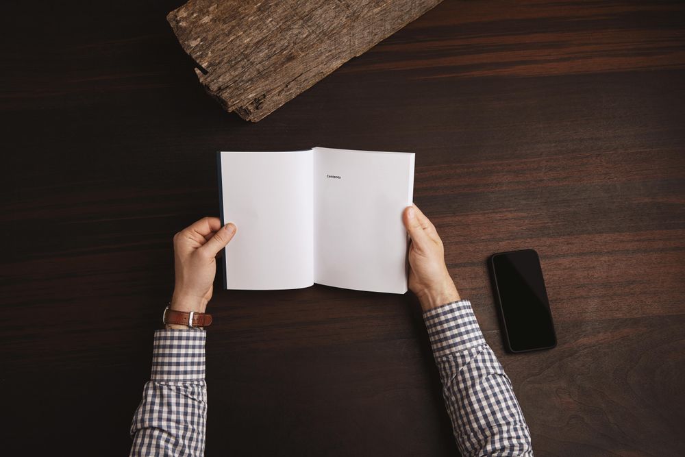 Arms in plaid shirt and watch holding open book with cell phone on table