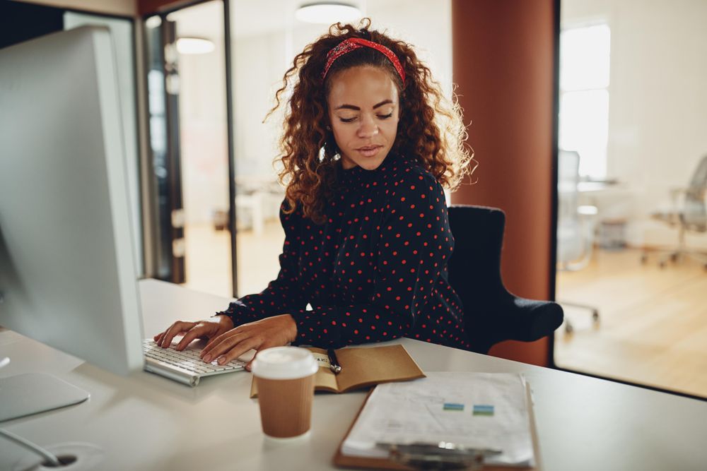 Woman with curly hair checking her notes and a coffee at computer at work