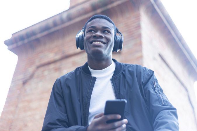 Smiling Black man outside, wearing headphones and holding phone