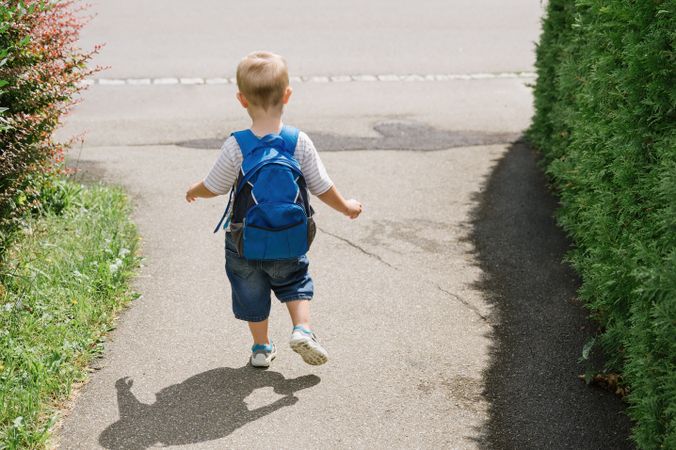 Unrecognizable little kid boy with backpack walking along road in sunlight