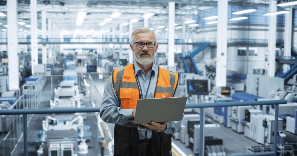 Portrait of Engineer Wearing Glasses, Using Laptop Computer and Looking at Camera. Technician in High-VIsibility Vest Working in a Modern Technology Factory