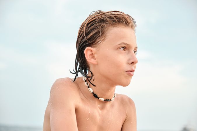 Shirtless boy wearing shell necklace looking away at beach