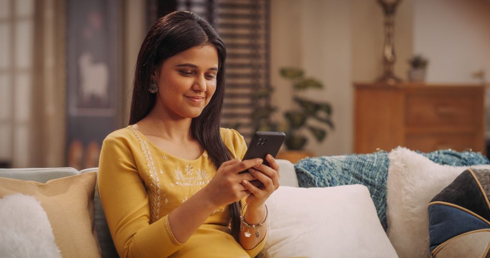 Young Indian Female Using Smartphone Sitting on a Couch in Cozy Home: Staying Connected with Friends, Browsing the Latest Trends