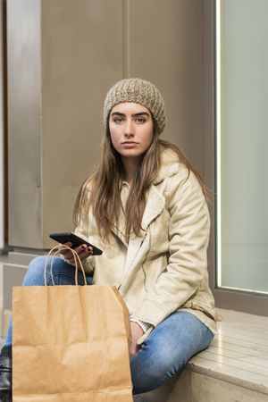 Woman sitting outdoors while using a phone with shopping bag