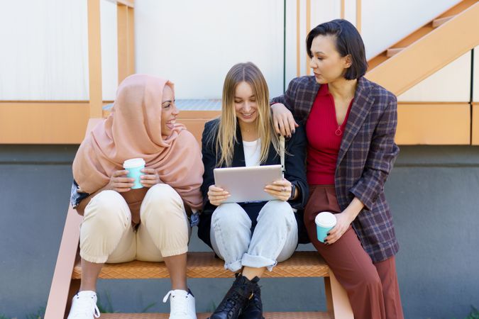Female friends sitting on outdoor staircase with coffee and digital tablet