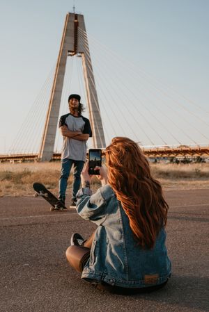 Woman siting on road and taking photo of boyfriend on smartphone