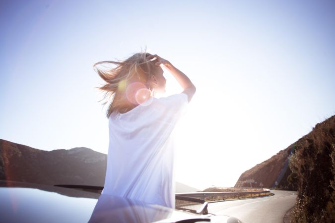 Woman standing out through a vehicle’s sunroof running her hands through her hair on bright day