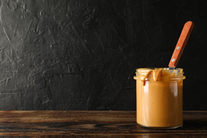 Glass jar with peanut butter and spoon on wooden table against dark background, space for text