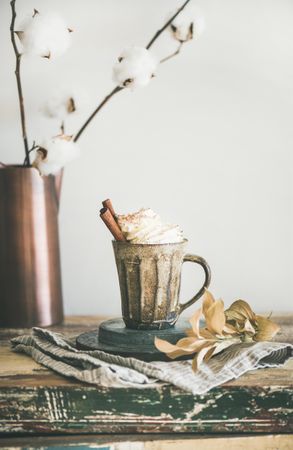 Mug of warm drink topped with whipped cream, on light background with dried cotton, copy space