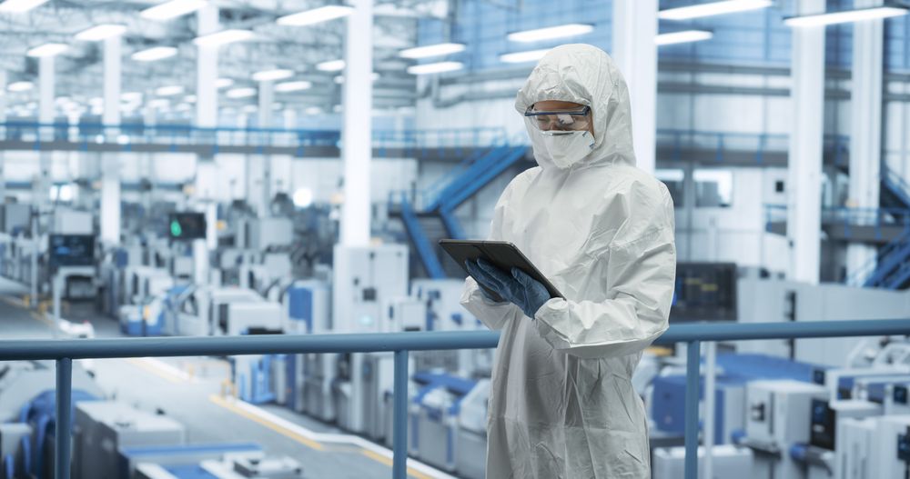 Industrial Scientist Wearing Disposable Protective Suit, Mask and Goggles at a Production Plant. Female Using Tablet Computer, Analyzing Potential Work Hazards and Contamination at a Workplace