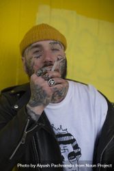 Punk hipster man wearing beanie with multiple facial tattoos smoking cigarette against yellow wall 48BKJ0