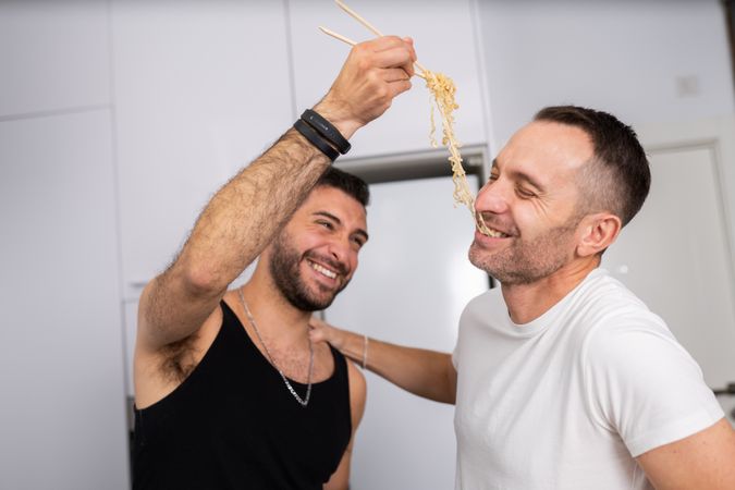 Laughing male couple playfully tasting noodles as they prepare meal