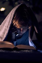 Boy reading reading book in darkness under blanket