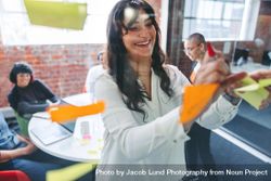 Smiling businesswoman sticking adhesive notes to a glass wall with her colleagues in the background 5zVeo5