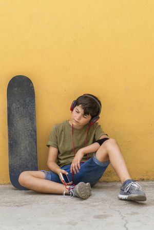 Tired teenager sitting on ground leaning on a yellow wall while holding a mobile phone