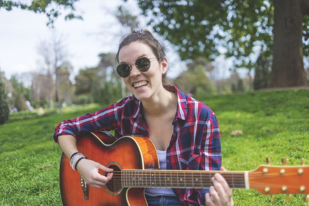 Front view of a young hipster woman wearing sunglasses, sitting on grass in a park while enjoying playing guitar