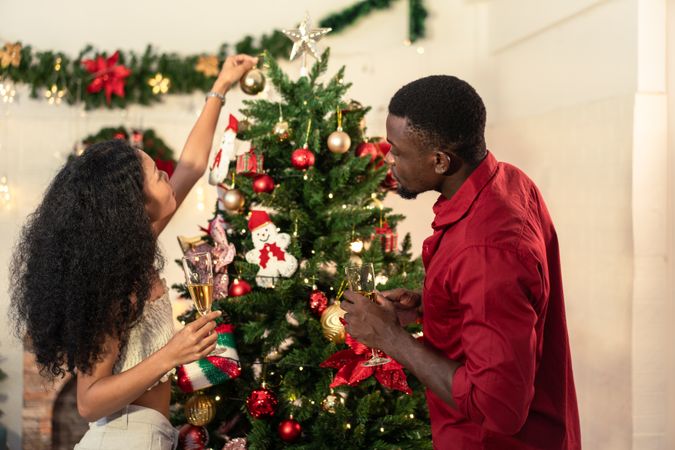 A Black couple decorates a Christmas tree while holding champagne glasses
