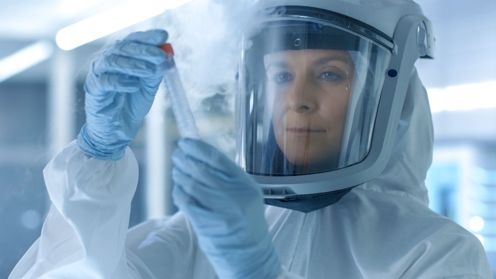 Medical Virology Research Scientist in a Hazmat Suit with Mask, She Inspects Test Tube with Isolated Virus String from Refrigerator Box.