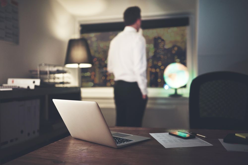 Shot of laptop and cell phone on desk with pensive man in shirt and tie in the background