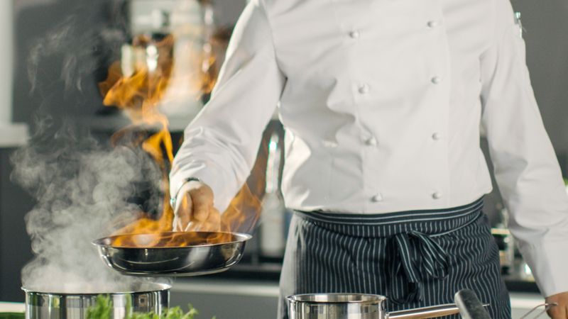 Close-up Shot of Fireing Oil on a Pan. Flambe Style Cooking. Cook Works in a Modern Kitchen with Lots of Ingredients are Around.