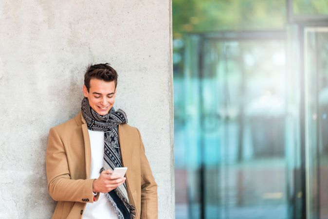 Smiling man leaning on wall outside texting and wearing a camel coat 