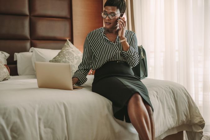 Woman in modern hotel room sitting on bed using laptop and talking over mobile phone