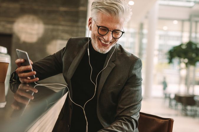 Smiling mature male in business suit sitting at cafe in earphones and holding a mobile phone