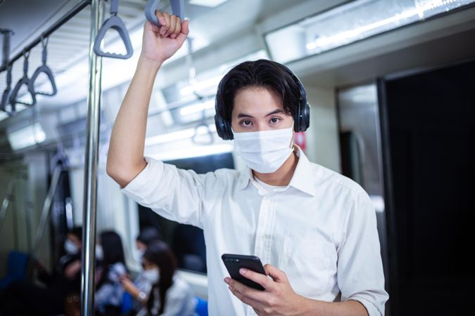 Man in facemask and headphones looking up from smartphone in metro car