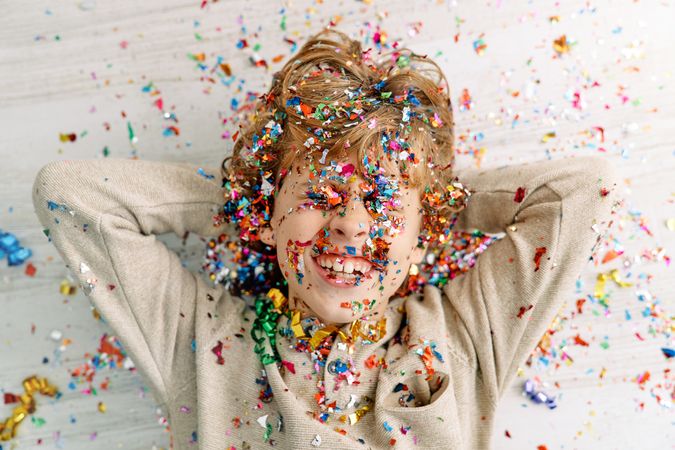 Happy boy covered with colorful confetti lying on floor